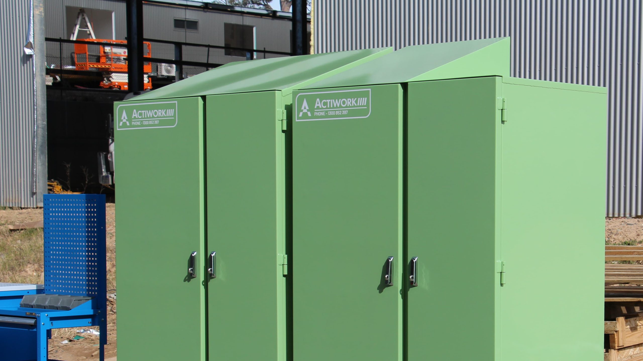 The image shows two large, green, industrial storage lockers placed on wooden pallets outdoors. The lockers have secure doors with handles and are labeled with "ACTIWORK." There is also a blue mobile tool cart with drawers and shelves next to the lockers.
