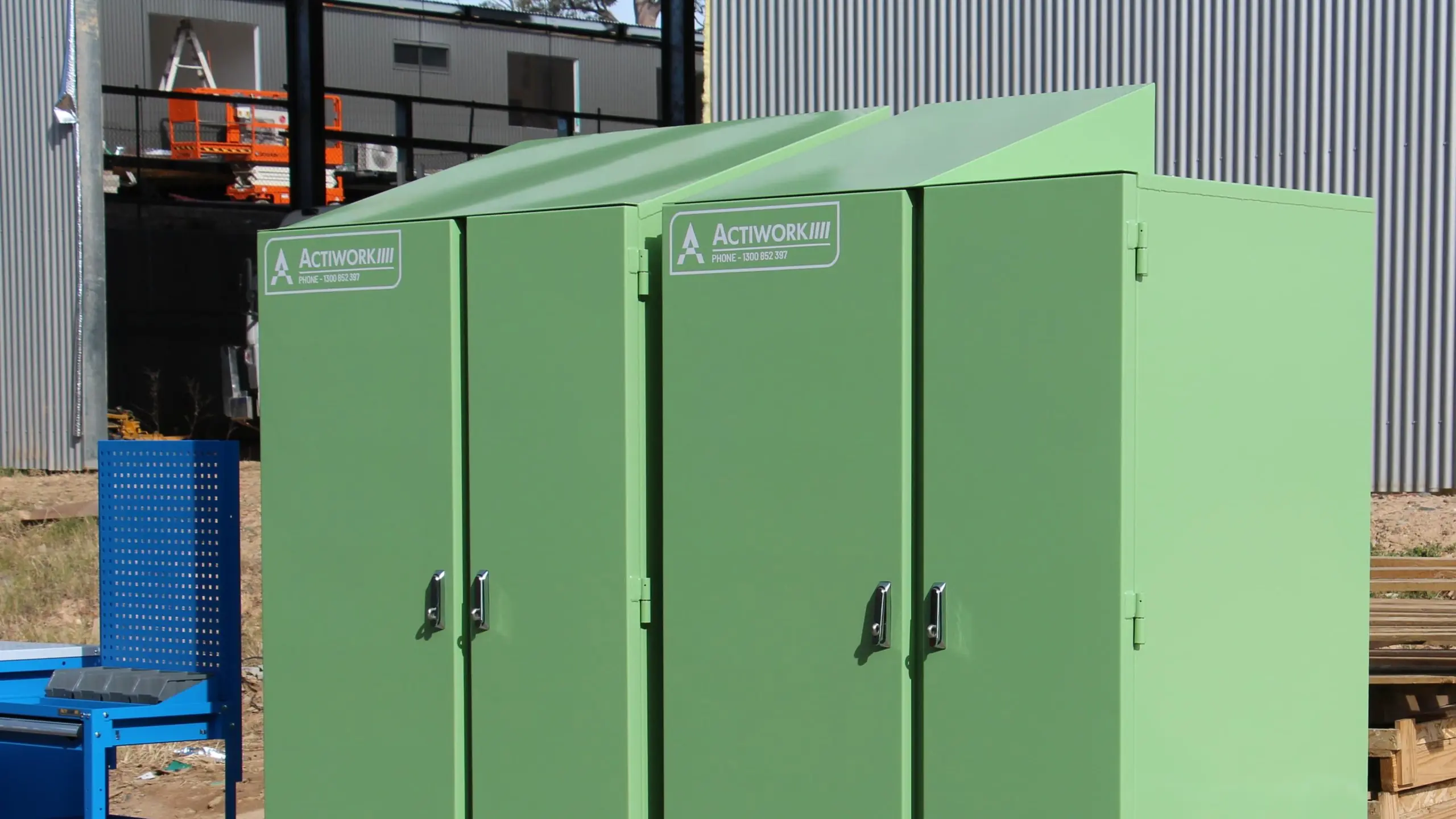 The image shows two large, green, industrial storage lockers placed on wooden pallets outdoors. The lockers have secure doors with handles and are labeled with "ACTIWORK." There is also a blue mobile tool cart with drawers and shelves next to the lockers.